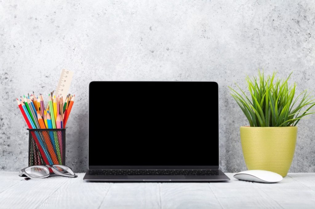 An open laptop sits on a desk with a pen pot filled with pencils and eyeglasses on the left and a potted pot on the left